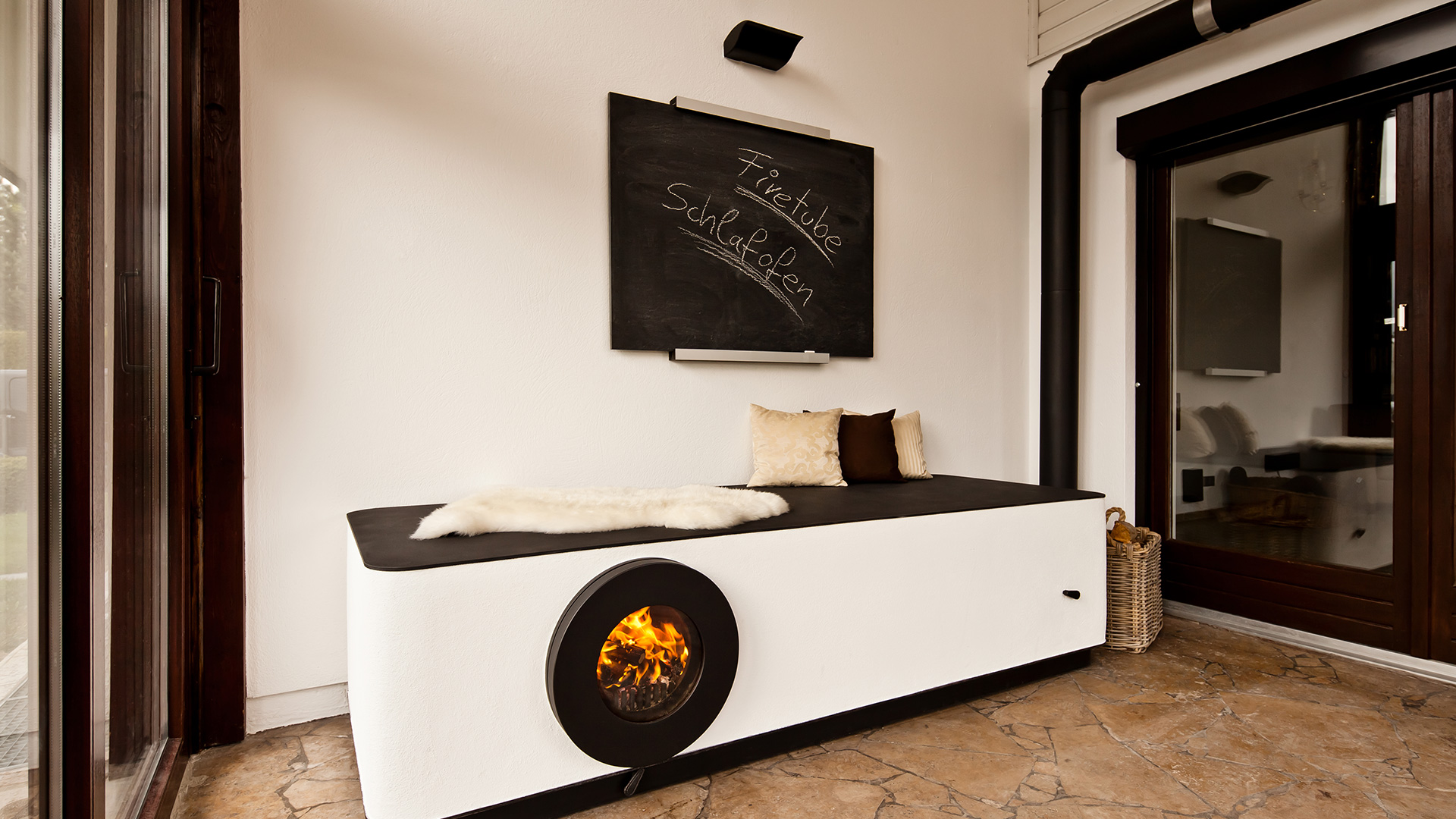 Firetube Ofen in Liegekonstruktion integriert mit sichtbarem Feuer; darauf Kissen und Fell, darüber Tafel mit Text „firetube Schlafofen“.