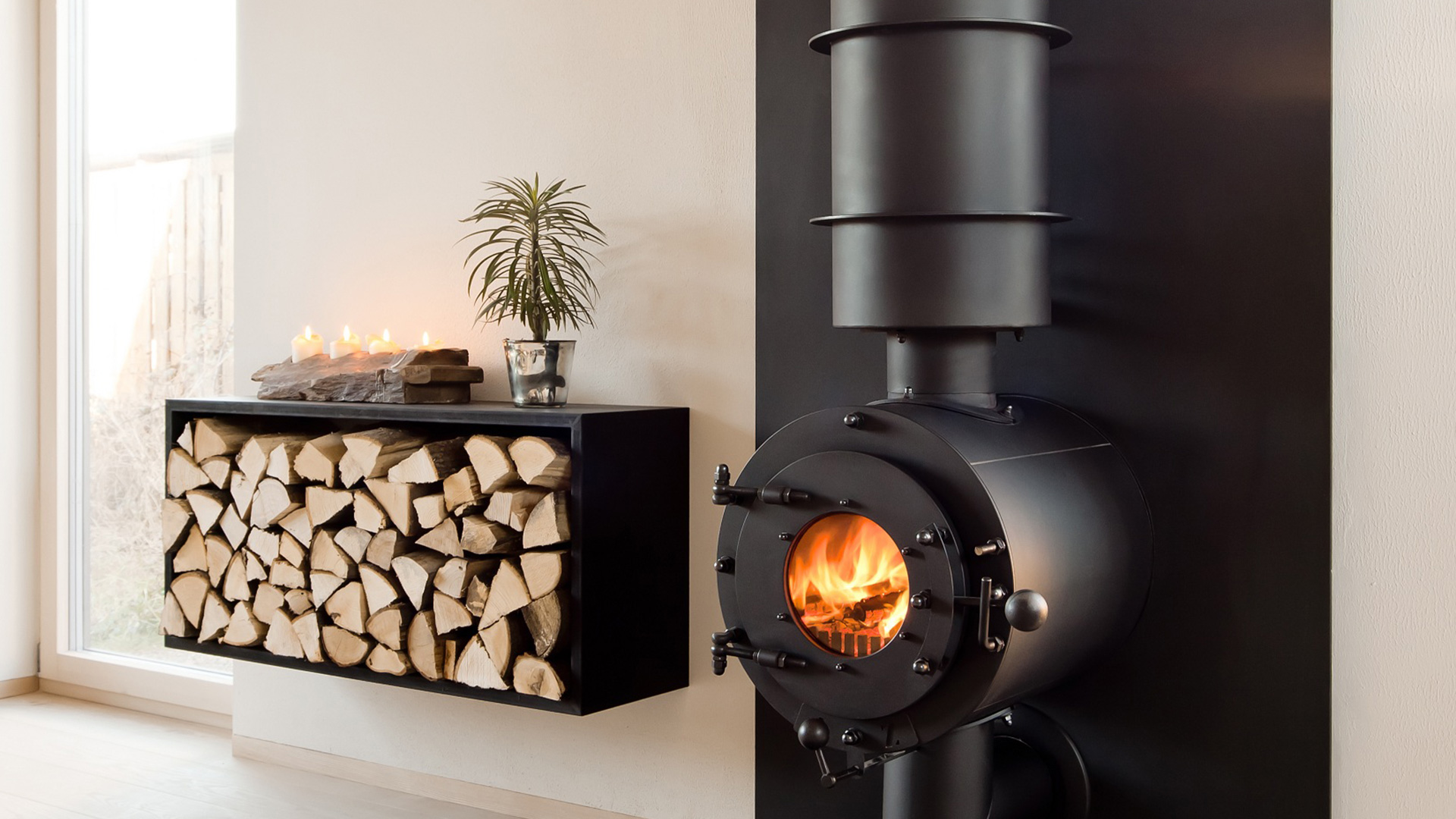 Schwarzer firetube Ofen mit sichtbarem Feuer an einer Wand; daneben Regal mit Holzscheiten und dekorativen Elementen.