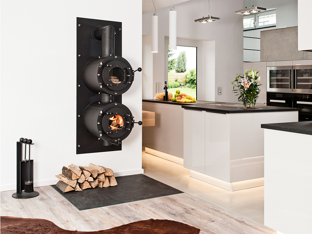 Schwarzer firetube Ofen in Wohnwand integriert, mit sichtbarem Feuer; darüber Garraum mit Brot, im Hintergrund eine Küche.