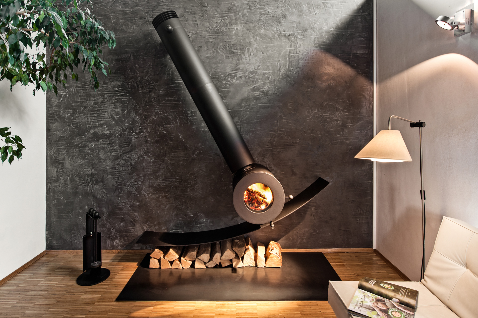Schräg an der Wand montierter firetube Individualofen mit sichtbarem Feuer; darunter Holzscheite, daneben eine Lampe in wohnlicher Atmosphäre.