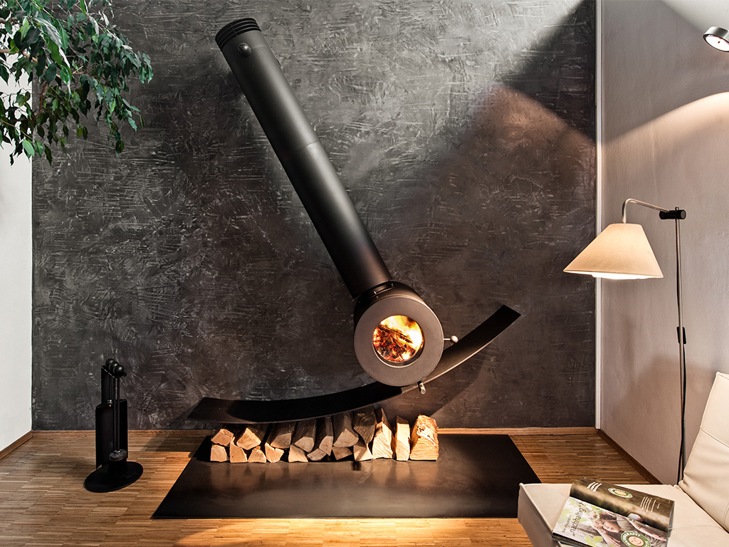 Schräg an der Wand montierter firetube Individualofen mit sichtbarem Feuer; darunter Holzscheite, daneben eine Lampe in wohnlicher Atmosphäre.