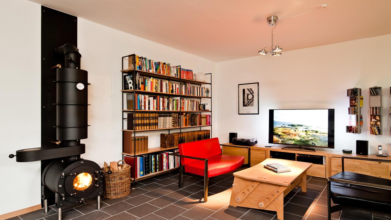 Schwarzer firetube Ofen mit sichtbarem Feuer und Hotplate an einer Wohnwand; im Hintergrund Bücherregal, Sitzmöglichkeiten und Fernseher.