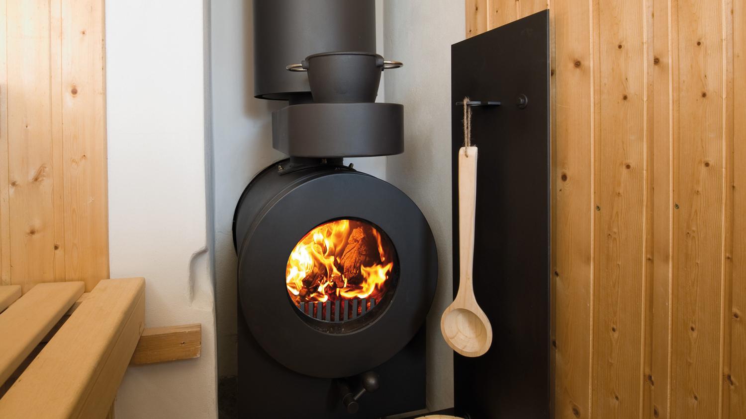 Schwarzer firetube Ofen in einer Sauna mit sichtbarem Feuer und Hotplate, zwischen Holzwand und Holzsitzbank.