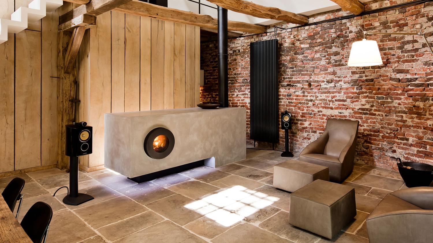 Wohnlich-rustikale Atmosphäre: grauer Steinblock mit integriertem schwarzen firetube Ofen und sichtbarem Feuer; dahinter Holzwand, rechts und links Lautsprecher, rechte Wand aus Backsteinen.