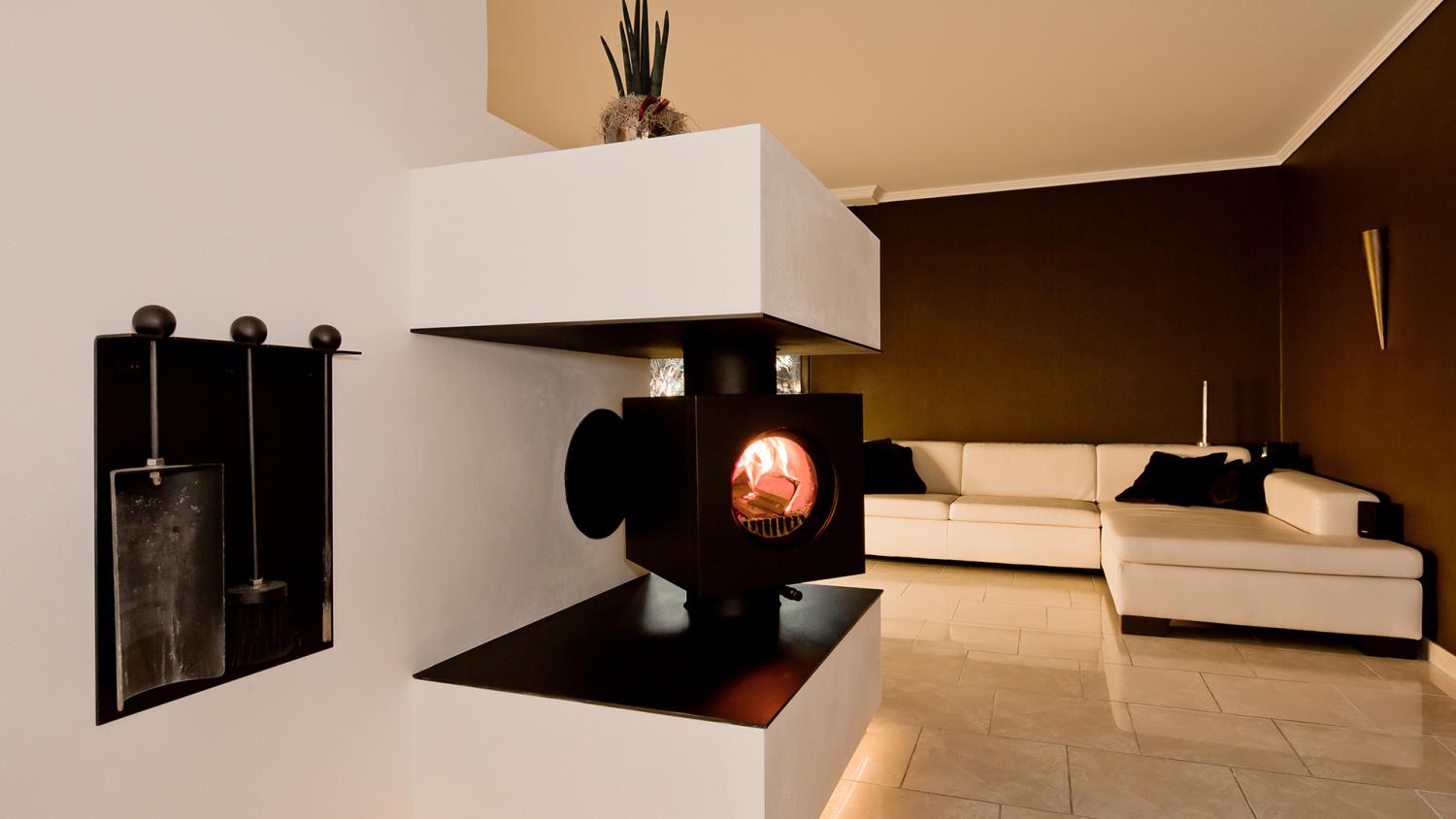 Schwarzer firetube Ofen mit sichtbarem Feuer, in eine raumtrennende Wand integriert; im Hintergrund ein Wohnzimmer.