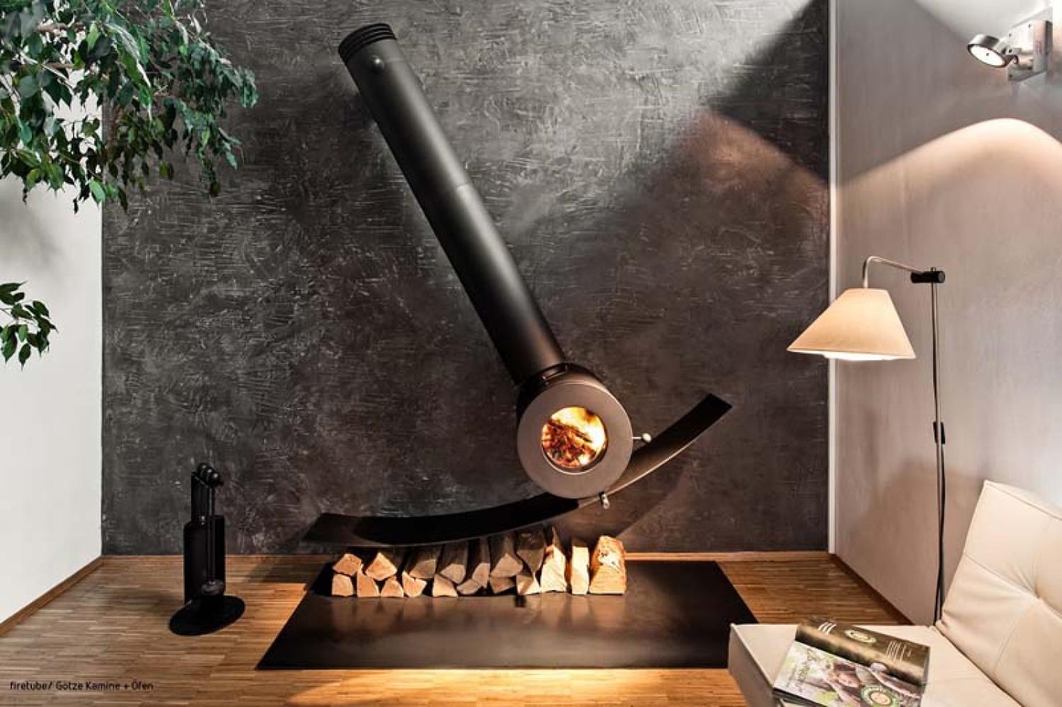 Schräg an der Wand montierter firetube Individualofen mit sichtbarem Feuer; darunter Holzscheite, daneben eine Lampe in wohnlicher Atmosphäre.