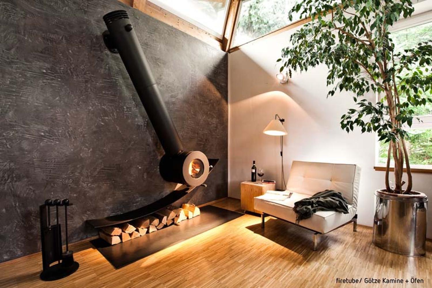 Schräg an der Wand montierter firetube Individualofen mit sichtbarem Feuer; darunter Holzscheite, daneben Lampe, Sitzecke und Pflanze in wohnlicher Atmosphäre.