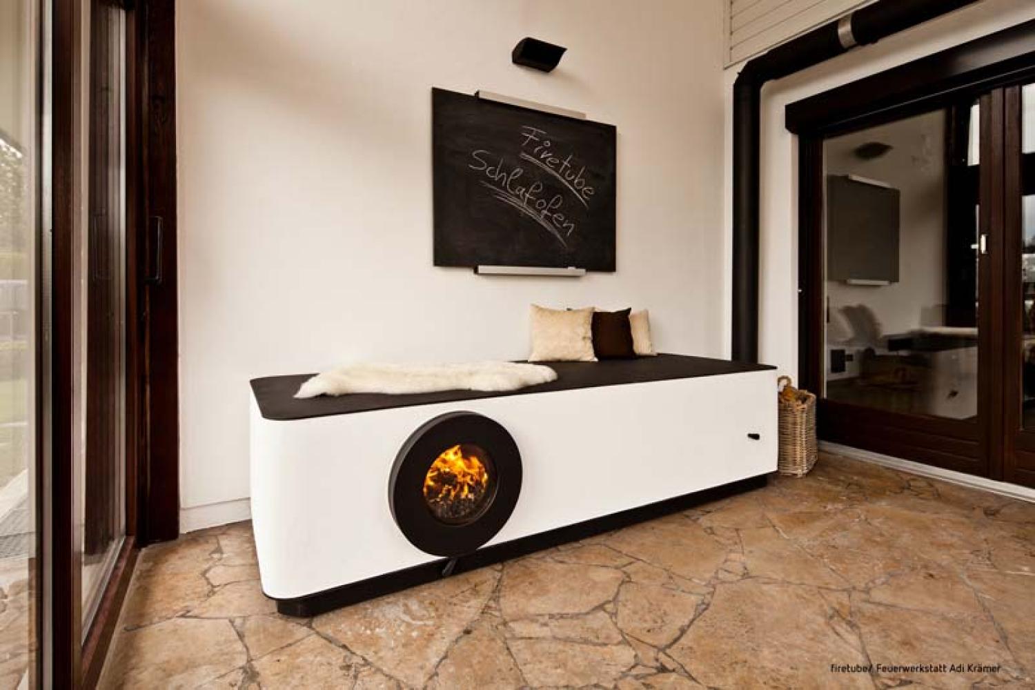 Firetube Ofen in Liegekonstruktion integriert mit sichtbarem Feuer; darauf Kissen und Fell, darüber Tafel mit Text „firetube Schlafofen“.