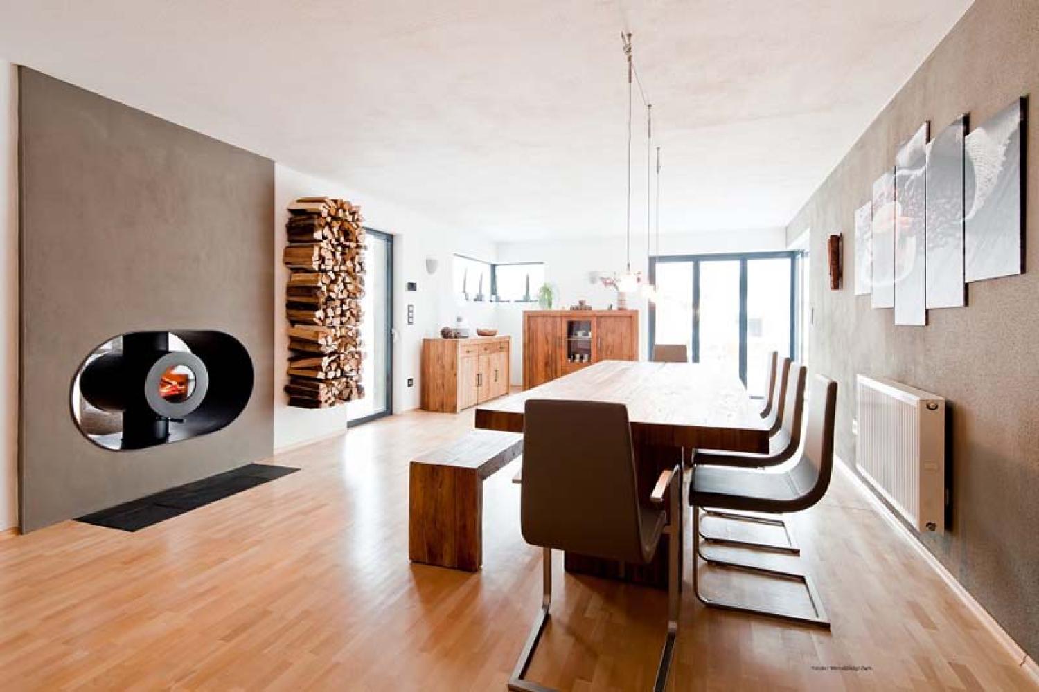 Schwarzer firetube Ofen mit sichtbarem Feuer, in Wohnwand integriert; daneben Holzscheite in der Wand, im Hintergrund Wohnzimmer mit Esstisch.