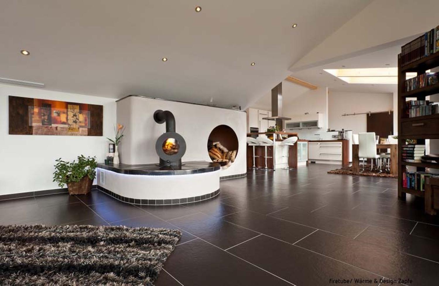 Großer Wohnraum mit schwarzem firetube Ofen und sichtbarem Feuer; davor Sitzmöglichkeit, daneben Nische mit Holzscheiten.