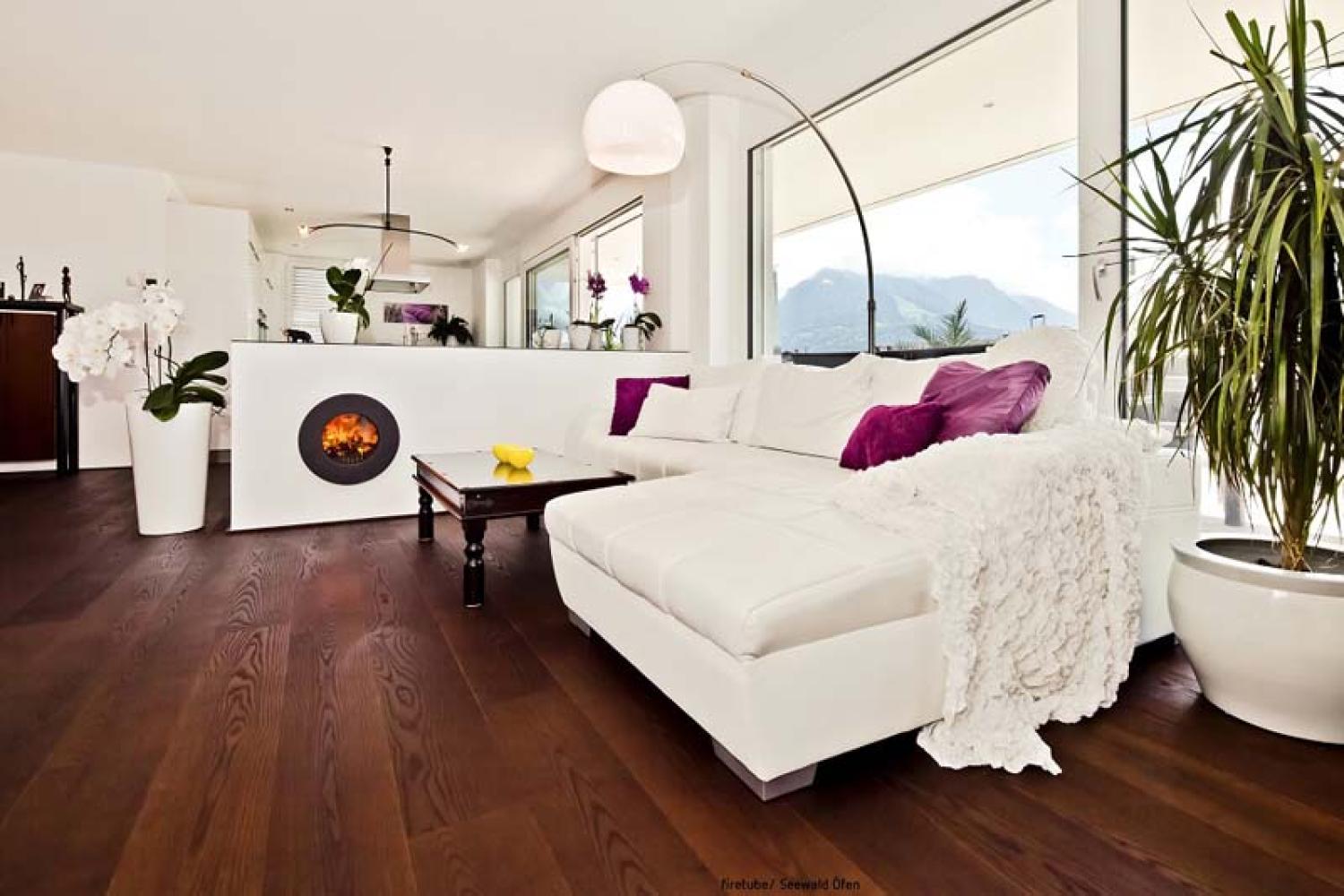 Weißes Sofa in einem Wohnzimmer; links davon trennende Wand mit integriertem schwarzem firetube Ofen und sichtbarem Feuer, im Hintergrund Fenster mit Ausblick.