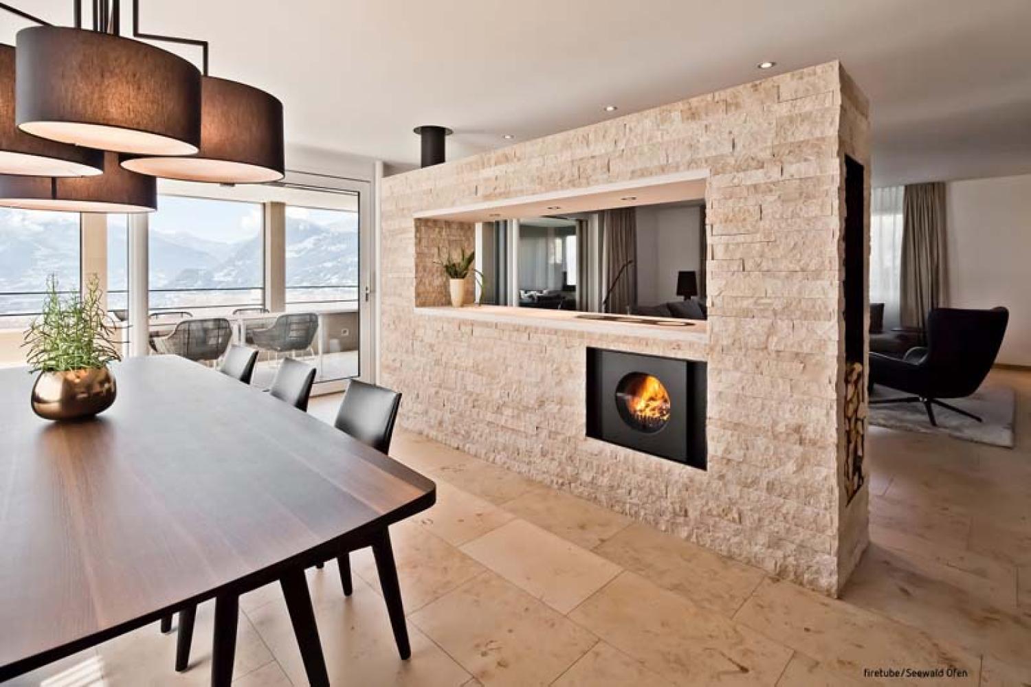 Wohnzimmer mit Esstisch und Fensterfront mit Aussicht; steinverkleidete Trennwand mit integriertem schwarzem firetube Ofen und sichtbarem Feuer.