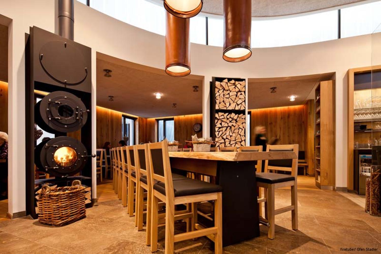 Lange Tafel in einem Gastronomieraum; links daneben schwarzer firetube Ofen mit sichtbarem Feuer, rechts gestapelte Holzscheite.