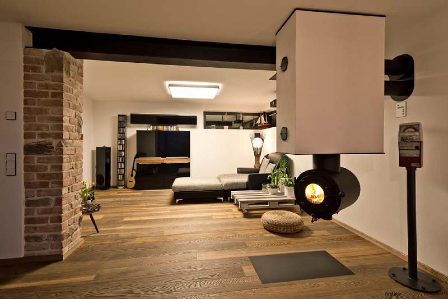 Modernes Wohnzimmer mit von der Decke hängendem schwarzen firetube Ofen und sichtbarem Feuer; im Hintergrund Möbel.