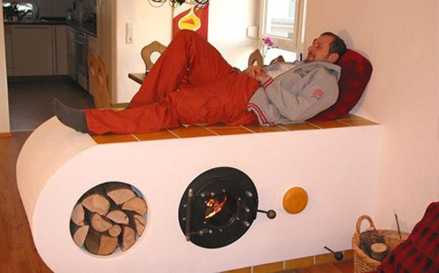 Mann liegt auf einer flachen Wand mit Liegefläche; darin ein firetube Ofen mit sichtbarem Feuer integriert, daneben Nische mit gestapelten Holzscheiten, im Hintergrund Küche.