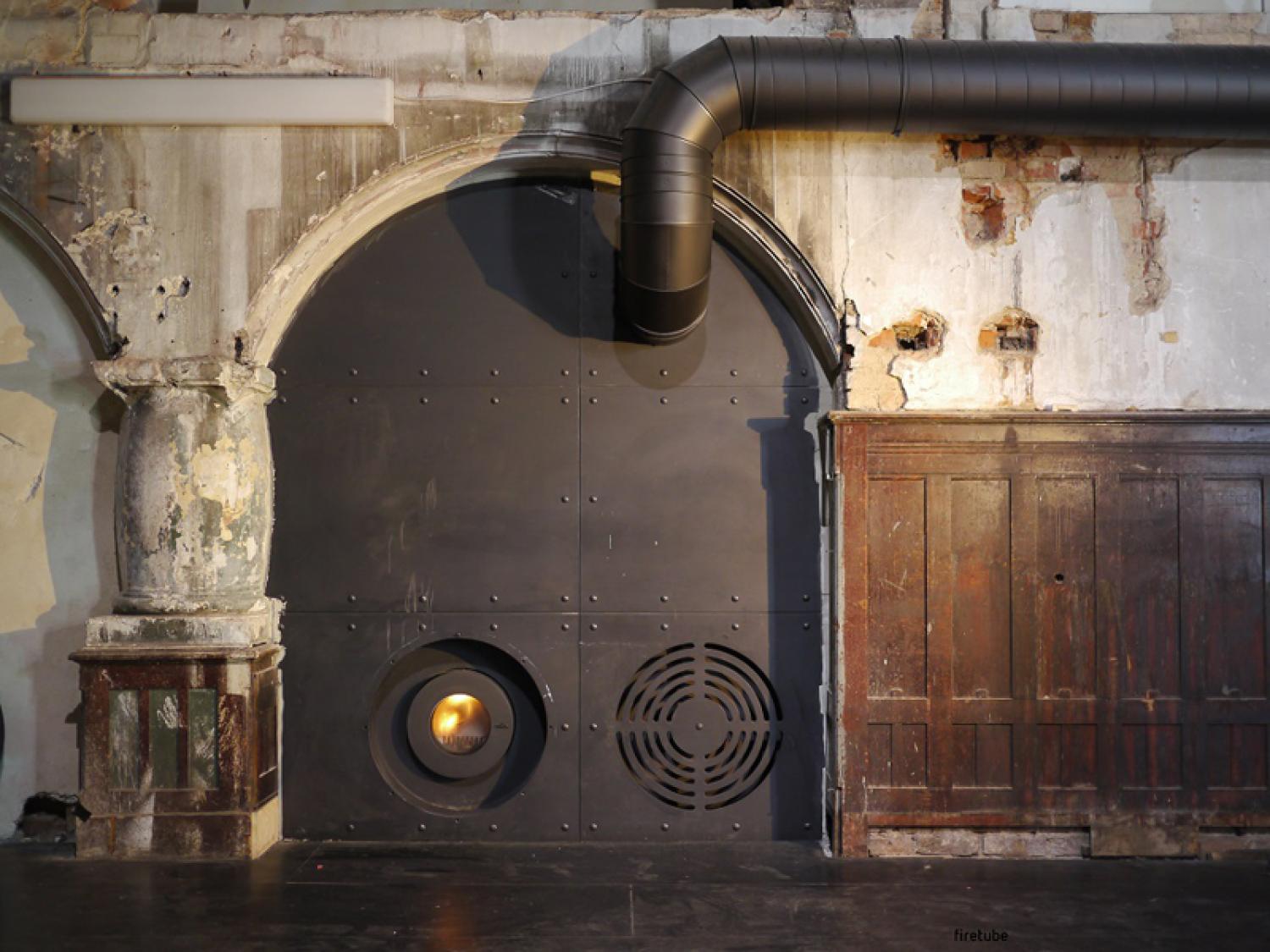 In eine rustikale Wand mit Türrahmen integrierter firetube Ofen aus dunklem Metall mit sichtbarem Feuer; unten links die Brennkammer, unten rechts eine Belüftungsöffnung.