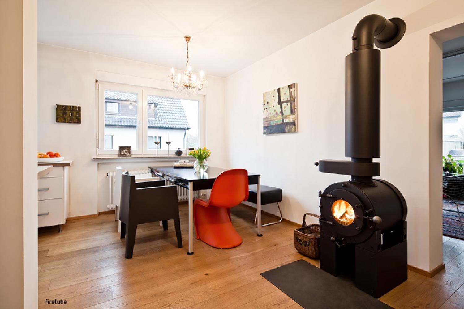 Gemütliches Zimmer mit Tisch am Fenster; am Zimmereingang ein freistehender schwarzer firetube Ofen mit sichtbarem Feuer und Hotplate.