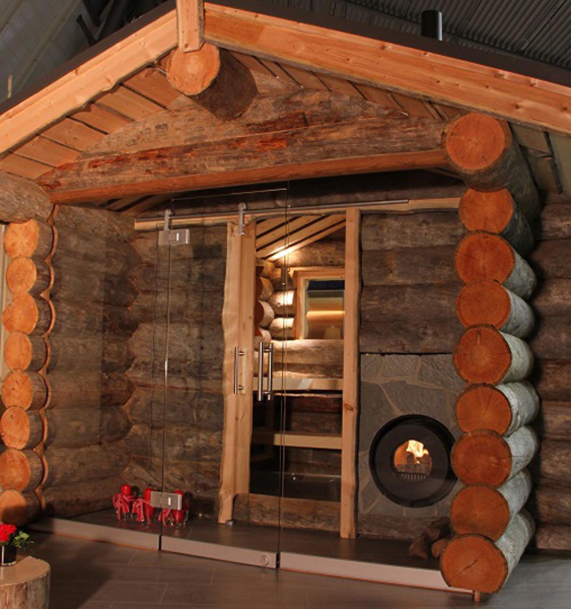 Holzhütte von außen mit Vorraum aus Glaswand und Glastür; darin ein firetube Ofen mit sichtbarem Feuer, in die Hauswand integriert.