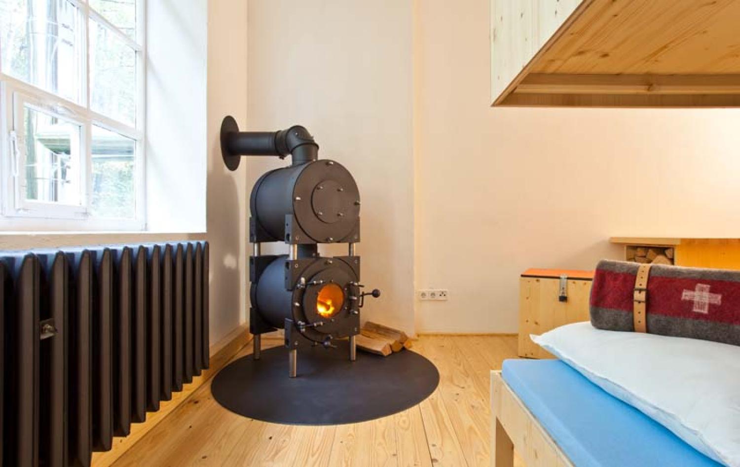 Schwarzer firetube Ofen mit sichtbarem Feuer und Garkammer an einer weißen Wand; links eine schwarze Heizung und Fenster.