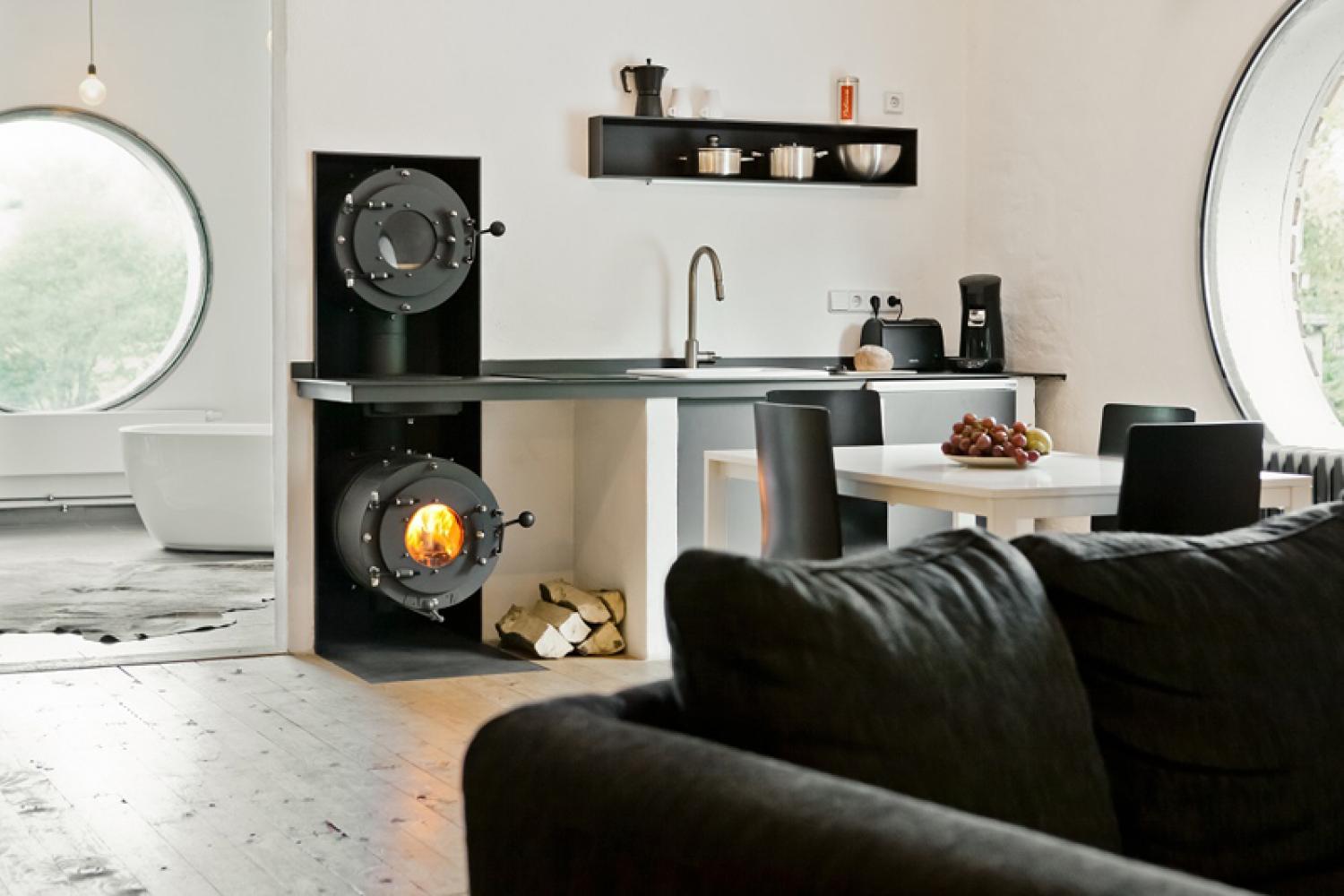 Modernes Wohnzimmer mit Küchenzeile an der Wand; links ein schwarzer firetube Ofen mit sichtbarem Feuer und darüber integriertem Garraum.