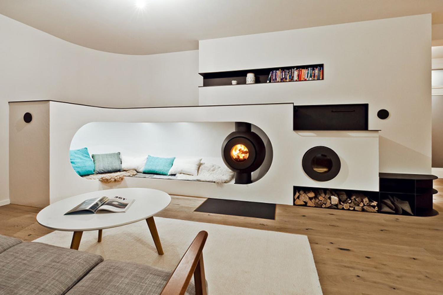 Schwarzer firetube Ofen mit sichtbarem Feuer, integriert in eine Sitzkuhle mit Kissen und Fell; umgeben von Wohnlandschaft.