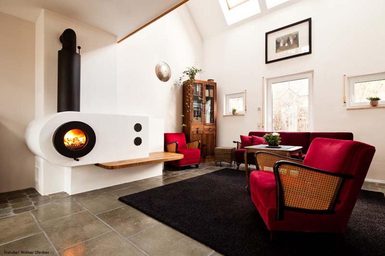 Schwarzer firetube Ofen mit sichtbarem Feuer, in Wohnwand integriert; im Hintergrund Wohnzimmer mit Sofa und Sessel.