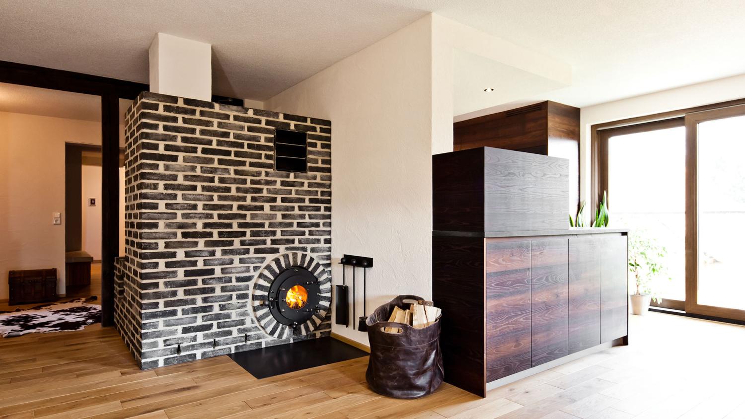 Schwarz-weiße Backsteinwand mit integriertem schwarzen firetube Ofen und sichtbarem Feuer; drumherum wohnliche Atmosphäre mit Regal und Fenster.