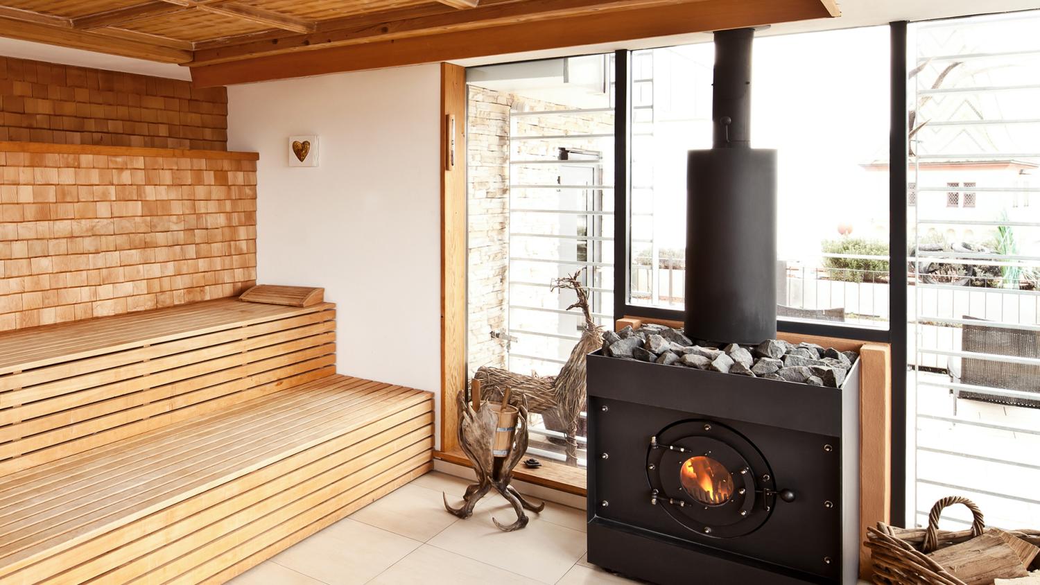 Firetube Saunaofen in einer Sauna mit Glaswand; daneben Holzdekoration, Korb mit Holzscheiten auf dem Boden und Sitzbänke aus Holz.