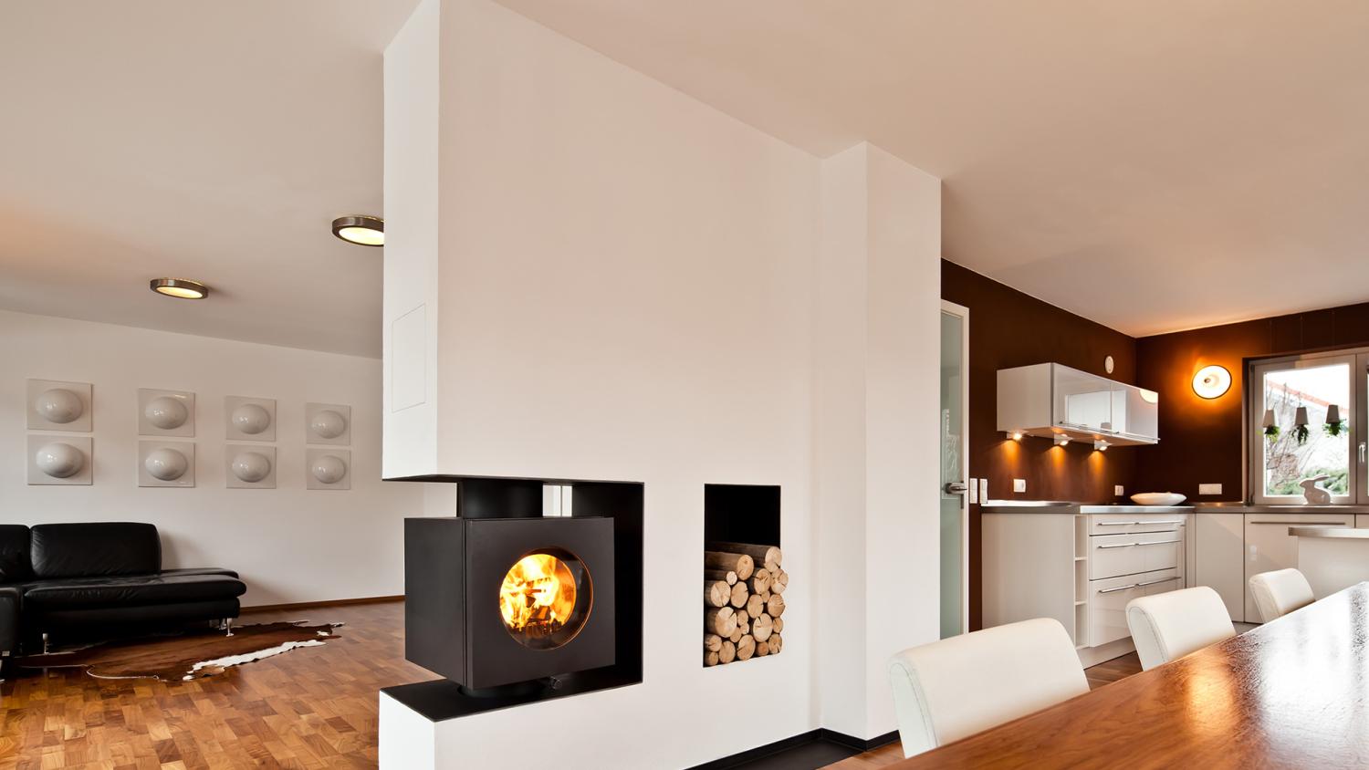 Rechteckiger firetube Ofen mit sichtbarem Feuer, integriert in eine Wohnwand mit Holzfach; Wohn- und Küchenbereich im Umfeld.