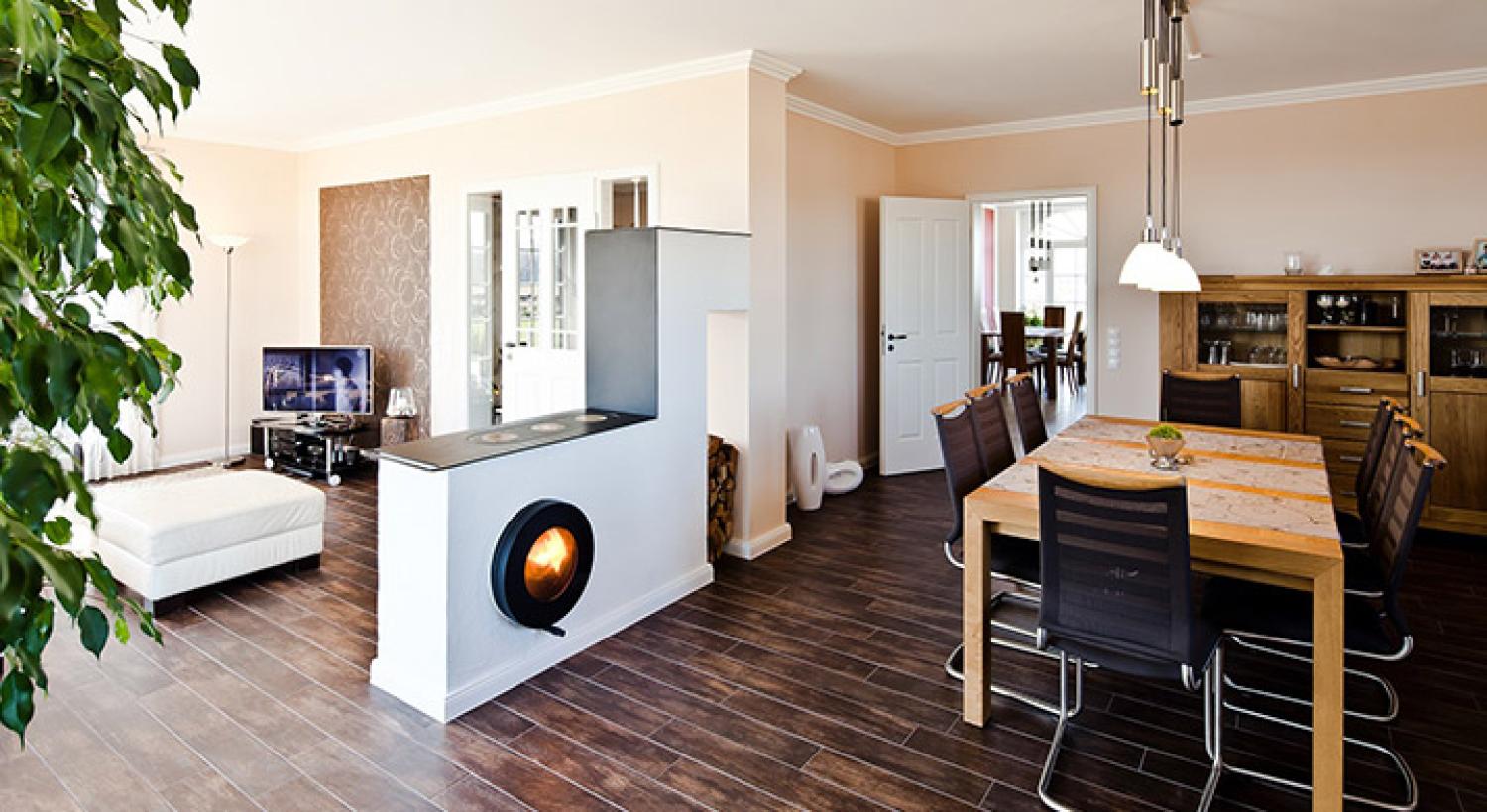 Firetube Ofen mit sichtbarem Feuer, in Küchenzeile integriert; darauf Kochplatten, dahinter gestapelte Holzscheite, drumherum Küchentisch und Wohnbereich.