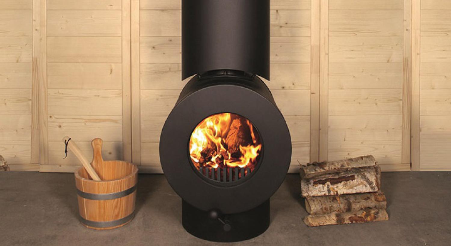 Schwarzer firetube Ofen mit sichtbarem Feuer vor Holzwand in Sauna-Umgebung; links Eimer, rechts Holzscheite.