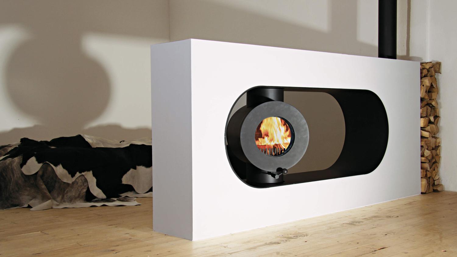 Schwarzer firetube Ofen mit brennendem Feuer, eingebaut in trennende flache Wand; dahinter Holzscheite.