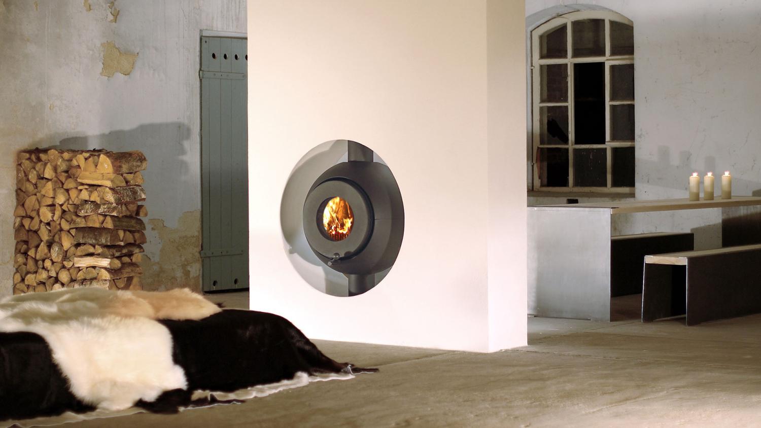 Schwarzer firetube Ofen mit sichtbarem Feuer, eingebaut in raumstehende Wand; dahinter Tisch und Holzscheite.