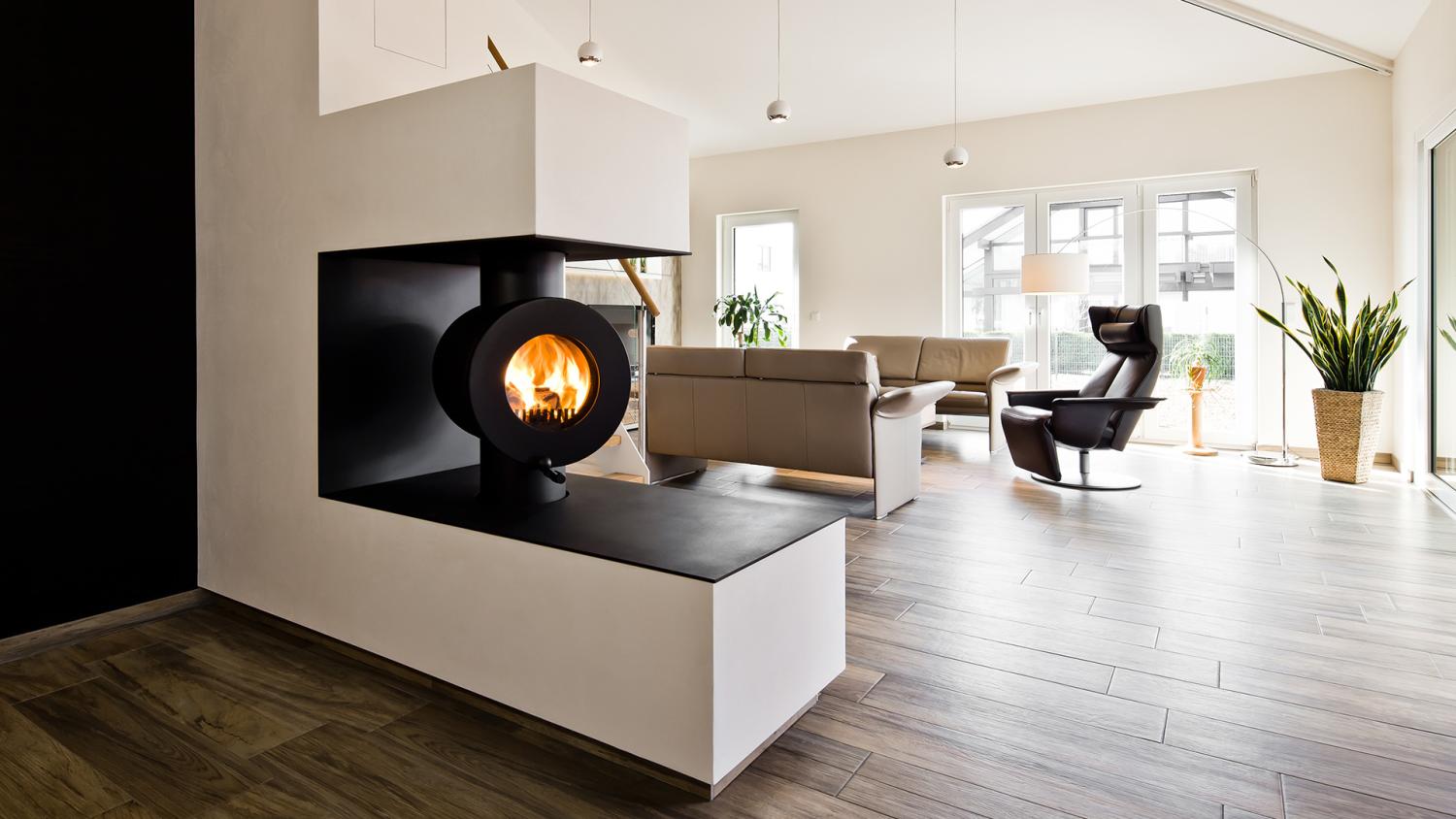 Schwarzer firetube Ofen mit sichtbarem Feuer, in eine raumtrennende Wand integriert; im Hintergrund ein Wohnzimmer.