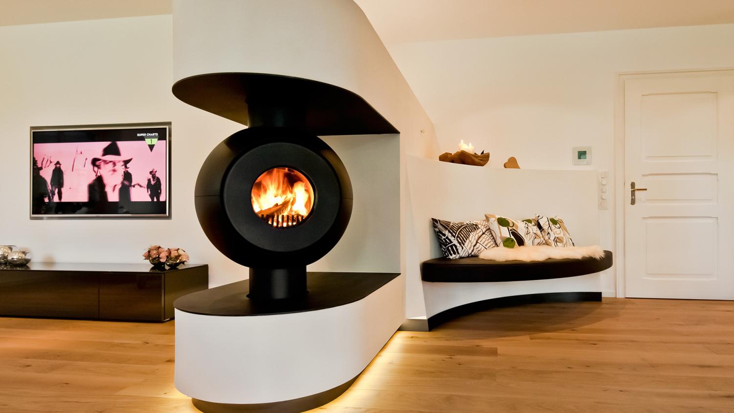 Schwarzer firetube Ofen mit sichtbarem Feuer, in eine raumtrennende Wand integriert; im Hintergrund ein Wohnzimmer mit Fernseher.