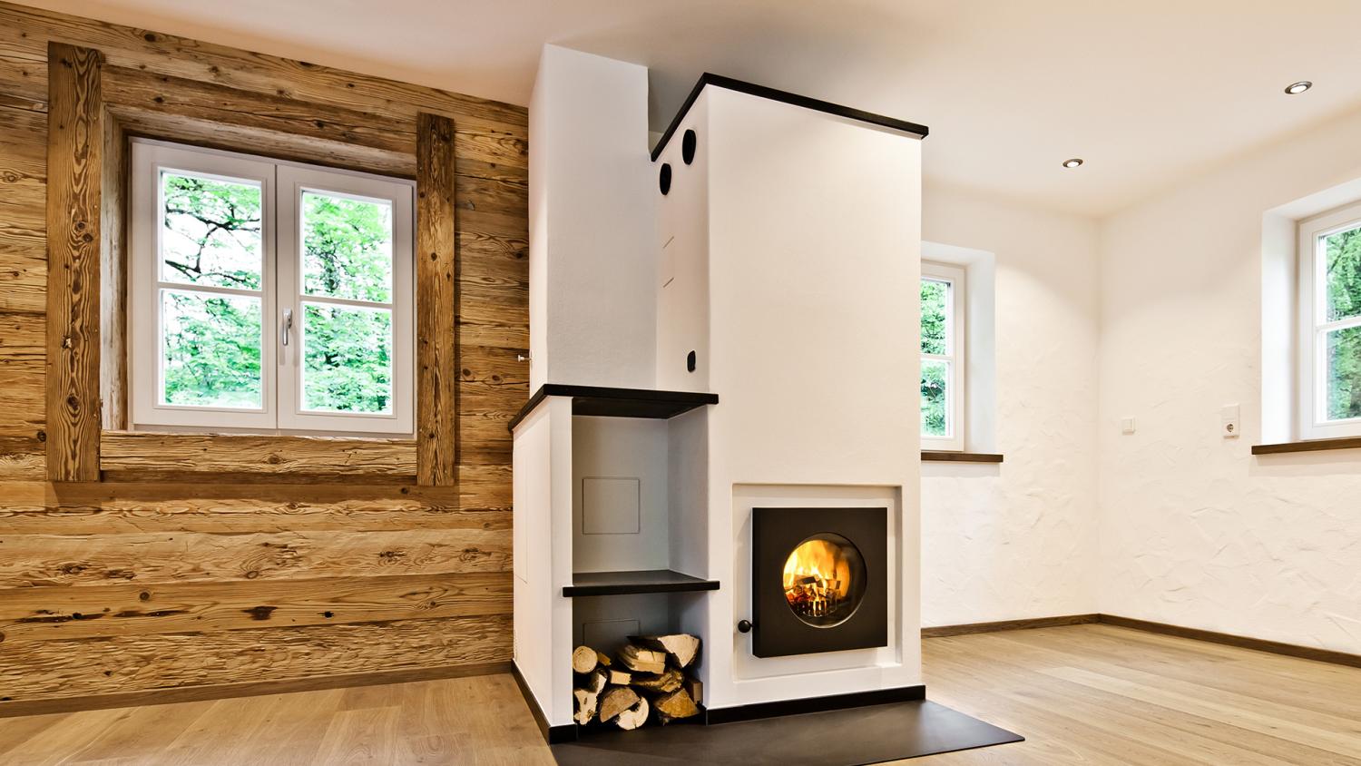 Firetube Ofen mit sichtbarem Feuer in einer trennenden Wand integriert; daneben Fach mit Holzscheiten, alles weiß verkleidet; linke Raumseite Holzwand, rechte Seite weiße Betonwand.