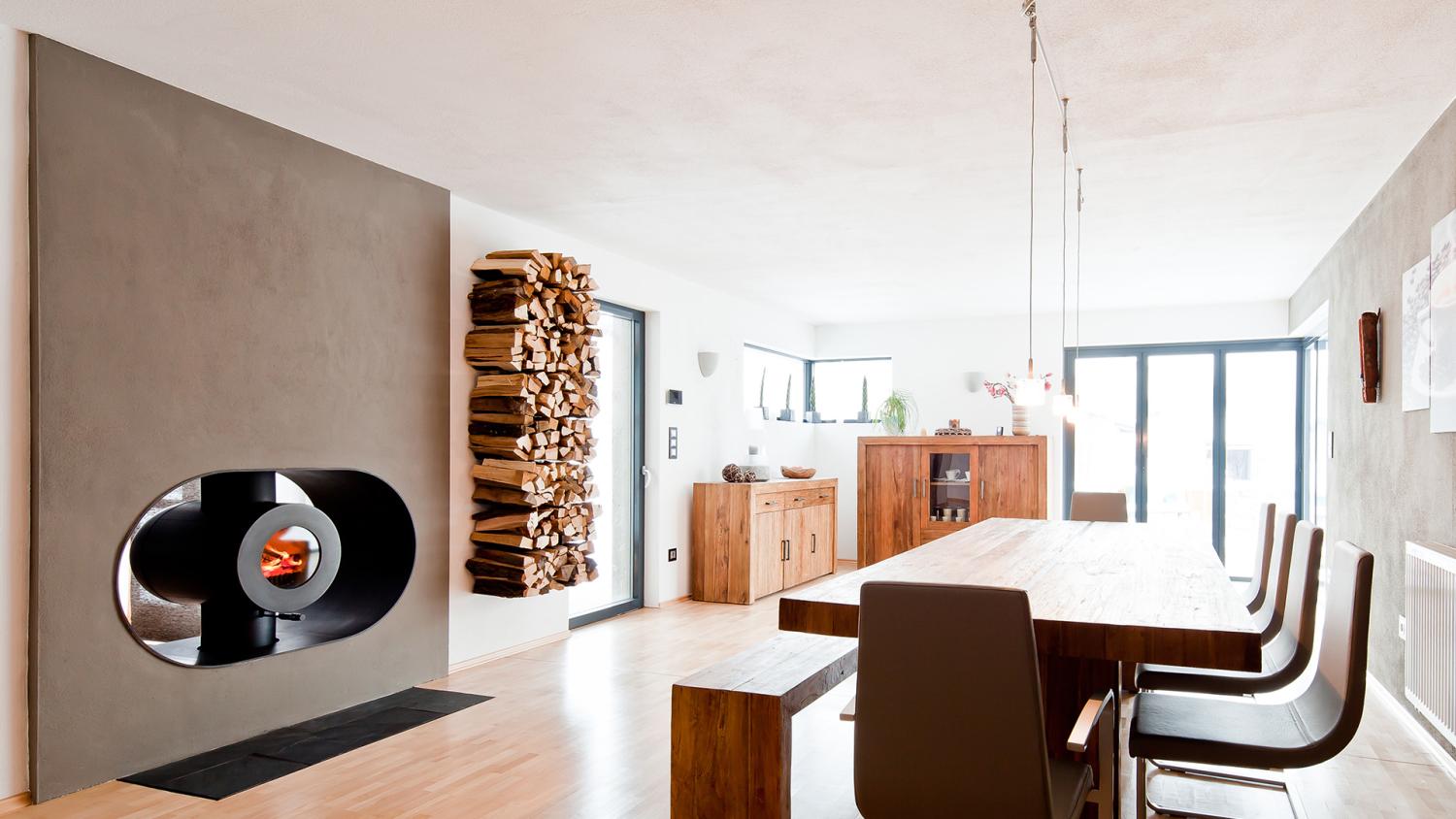 Schwarzer firetube Ofen mit sichtbarem Feuer, in Wohnwand integriert; daneben Holzscheite in der Wand, im Hintergrund Wohnzimmer mit Esstisch.