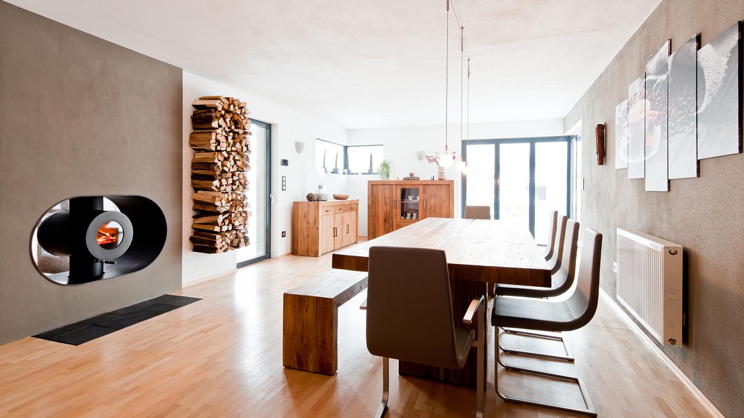 Schwarzer firetube Ofen mit sichtbarem Feuer, in Wohnwand integriert; daneben Holzscheite in der Wand, im Hintergrund Wohnzimmer mit Esstisch.