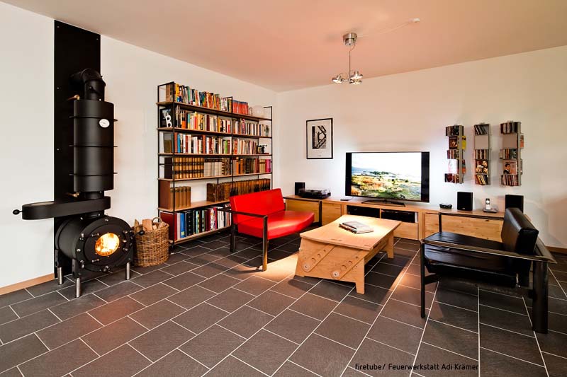 Schwarzer firetube Ofen mit sichtbarem Feuer und Hotplate an einer Wohnwand; im Hintergrund Bücherregal, Sitzmöglichkeiten und Fernseher.
