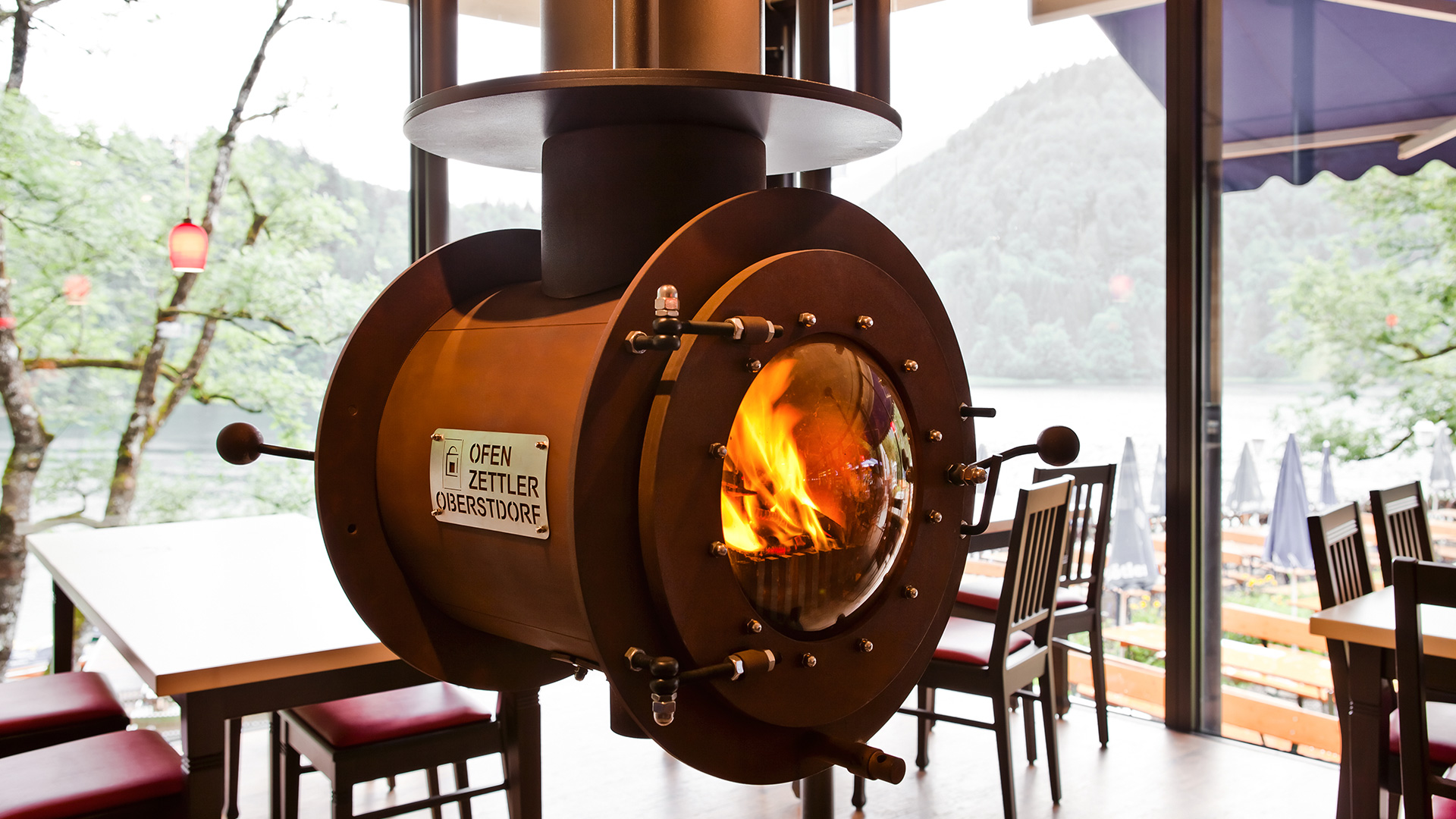 Von der Decke hängender schwarzer firetube Ofen mit sichtbarem Feuer in gastronomischer Umgebung; Tische im Hintergrund.