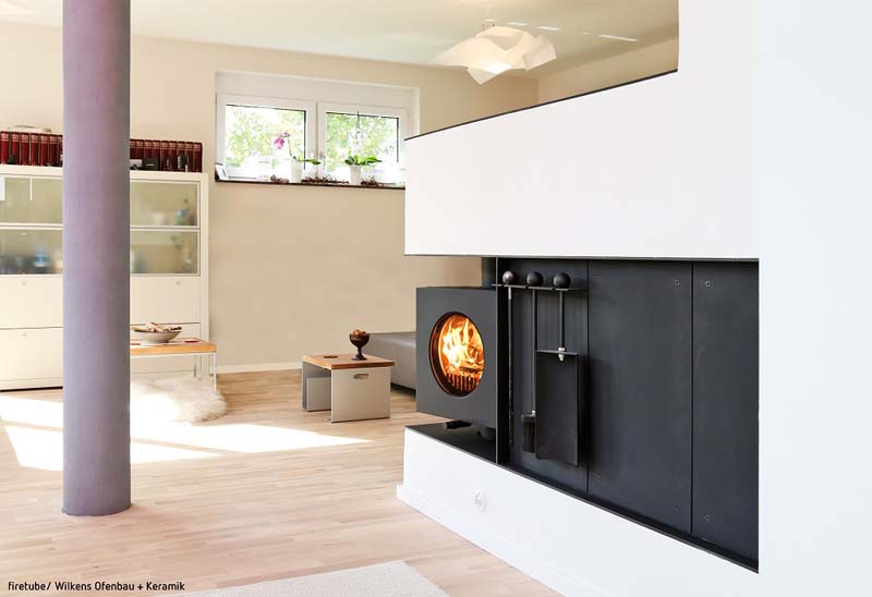Schwarzer firetube Ofen mit sichtbarem Feuer, integriert in eine Wohnwand; im Hintergrund ein wohnliches Zimmer mit Fenstern, daneben eine lilafarbene Säule.