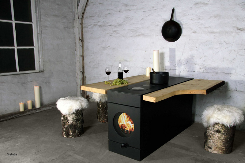firetube Ofen mit sichtbarem Feuer, integriert in eine Küchenzeile mit Wärmeplatten und Topf; Holzverlängerung mit Weingläsern, Trauben und Kerzen, rustikales Ambiente mit Holzhockern und Fell.