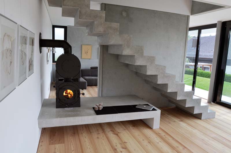 Schwarzer firetube Ofen mit sichtbarem Feuer und Garkammer über der Brennkammer, wandmontiert in modernem Wohnzimmer mit integrierter Sitzgelegenheit; daneben eine rustikale Betontreppe und Fenster im Hintergrund.