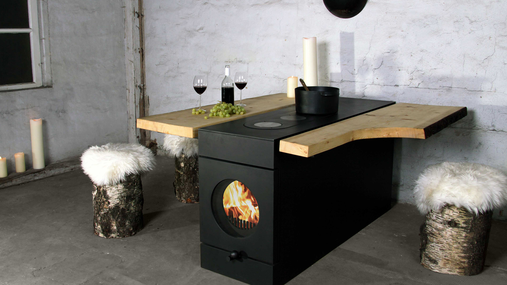 firetube Ofen mit sichtbarem Feuer, integriert in eine Küchenzeile mit Wärmeplatten und Topf; Holzverlängerung mit Weingläsern, Trauben und Kerzen, rustikales Ambiente mit Holzhockern und Fell.