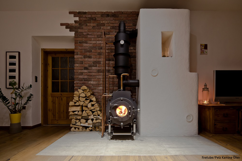 Schwarzer firetube Ofen mit sichtbarem Feuer in wohnlicher Atmosphäre; links daneben gestapelte Holzscheite.