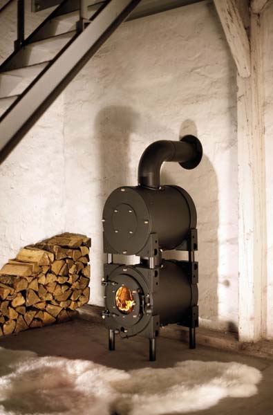 Schwarzer firetube Ofen mit sichtbarem Feuer und Garkammer an einer weißen Steinwand; links gestapelte Holzscheite, im Hintergrund eine Treppe.