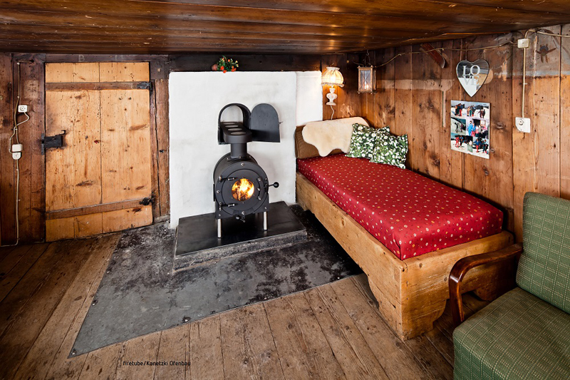 Gemütliches Zimmer mit Holzboden und Holzwänden, kleines Bett mit rotem Überzug; in der Mitte schwarzer firetube Ofen mit sichtbarem Feuer.