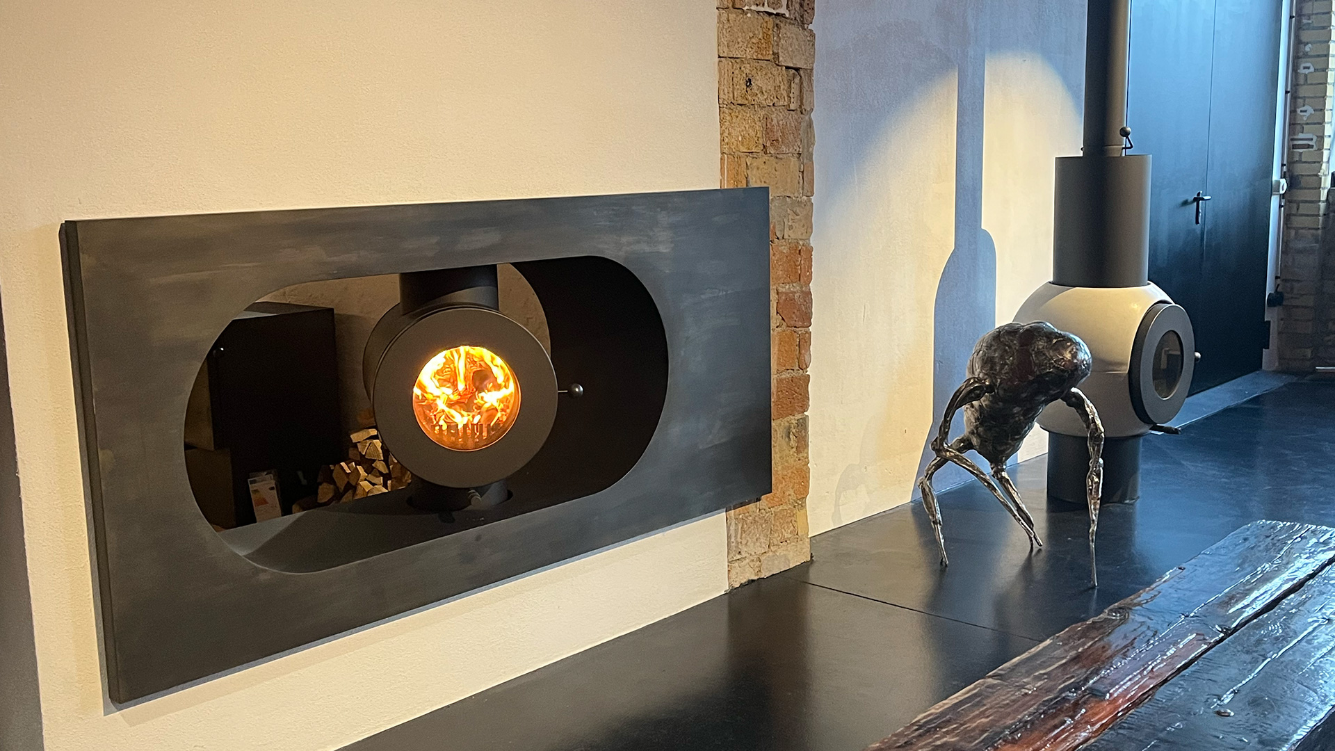 Schwarzer firetube Ofen mit sichtbarem Feuer, in eine Wand integriert; hinter dem Ofen Holzscheite, rechts auf dem Boden ein weiterer firetube Ofen.