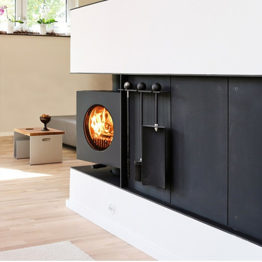 Schwarzer firetube Ofen mit sichtbarem Feuer, eingearbeitet in trennende Wohnwand; dahinter Ausschnitt eines Wohnzimmers.