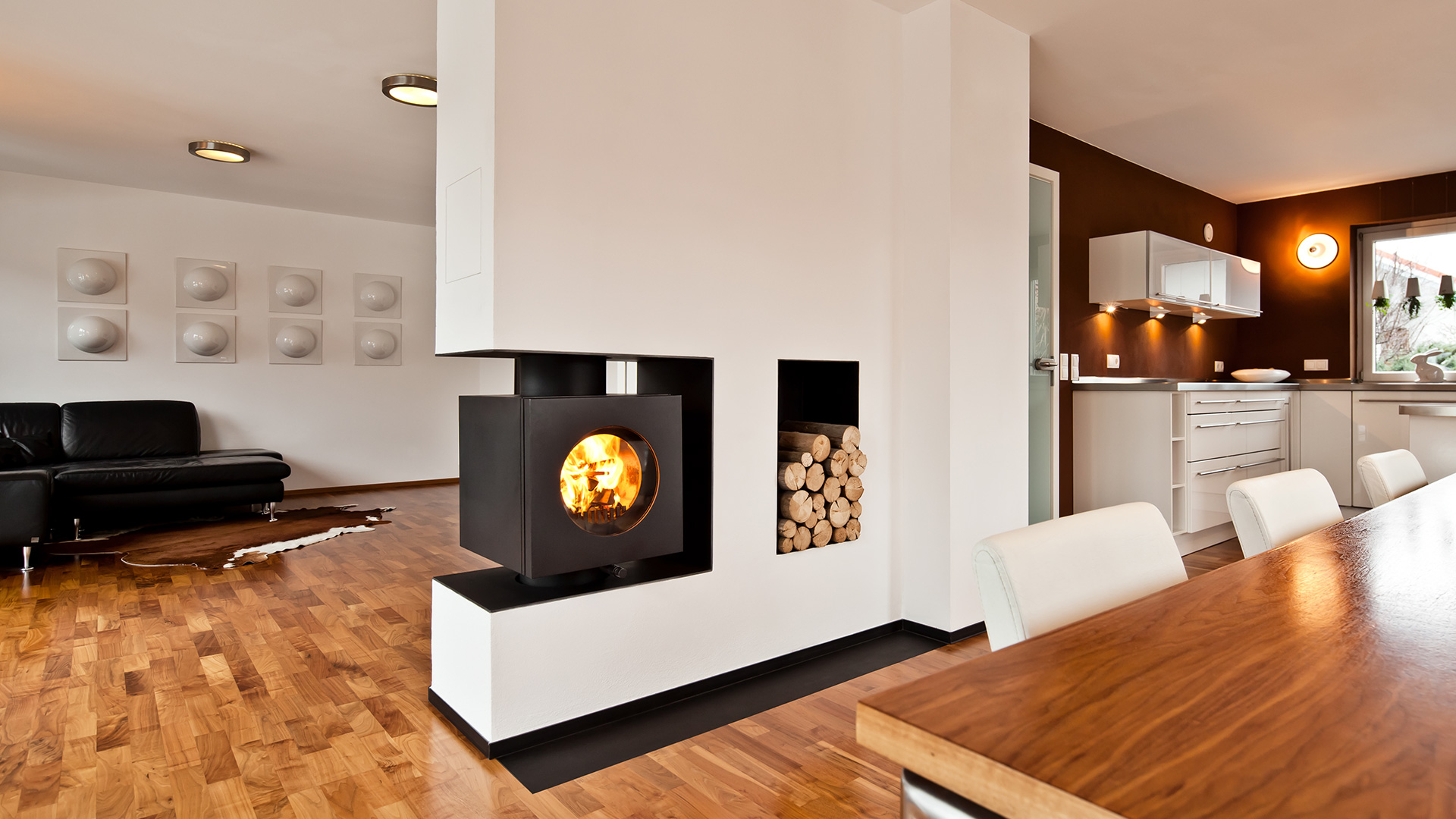 Rechteckiger firetube Ofen mit sichtbarem Feuer, integriert in eine Wohnwand mit Holzfach; Wohn- und Küchenbereich im Umfeld.